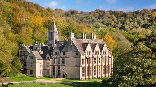 A view of Woodchester Mansion on a sunny day in Woodchester Park, surrounded by green and brown trees in autumn